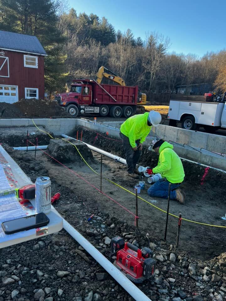 Construction workers in neon vests install pipes at a foundation site with a red dump truck and excavator.