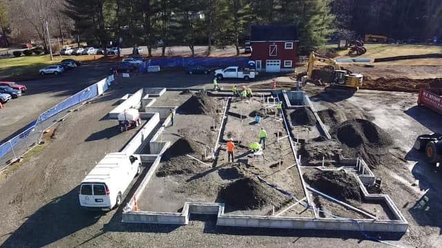Construction site: Workers building a foundation, dirt piles, white van, blue fencing, and red building in the background.