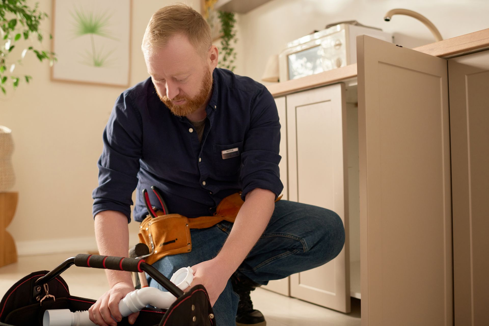 Plumber kneeling, working on plumbing under a kitchen sink. He wears a tool belt and examines a pipe.