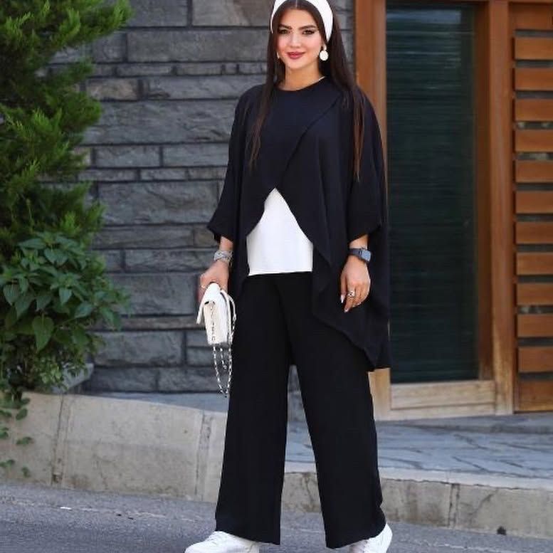 A woman wearing a black cardigan and black pants is standing in front of a building holding a white purse.