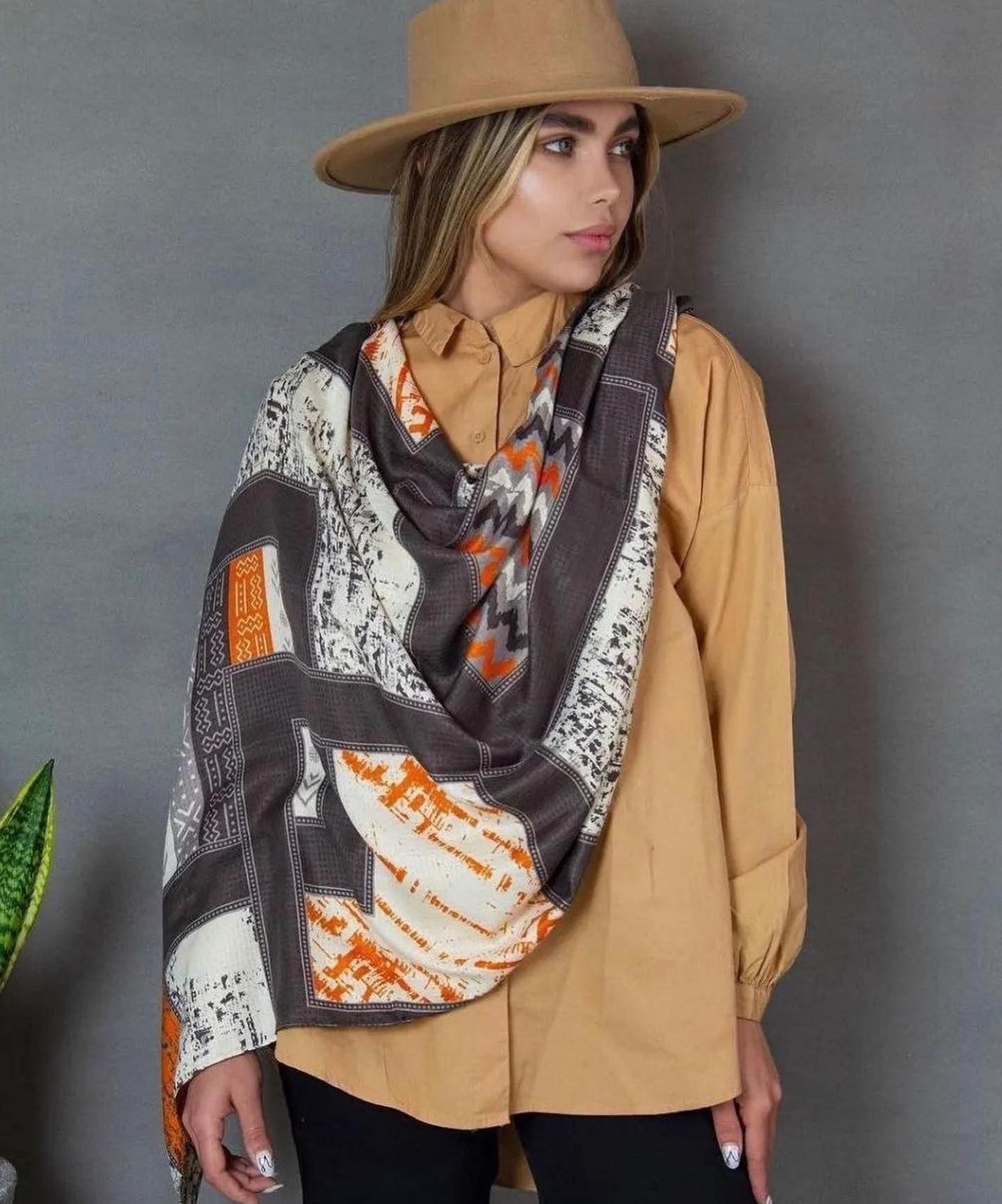 A woman wearing a hat and a scarf is standing in front of a plant.