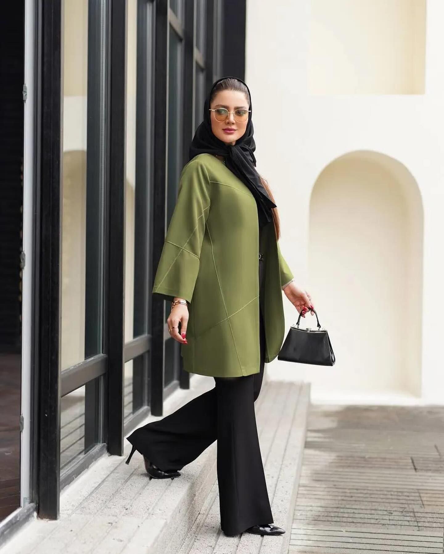 A woman wearing a green coat and black pants is holding a black purse.