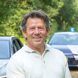 Een man in een wit overhemd staat lachend voor twee auto's.