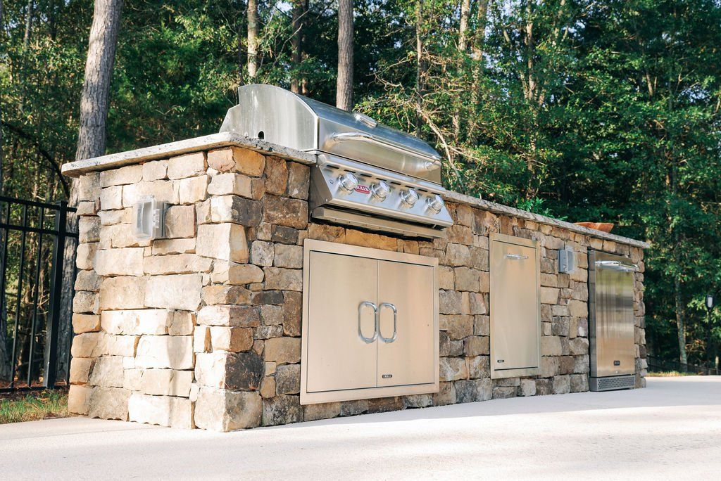 Stone outdoor barbecue grill built into a wall in a wooded area, with stainless steel accents.