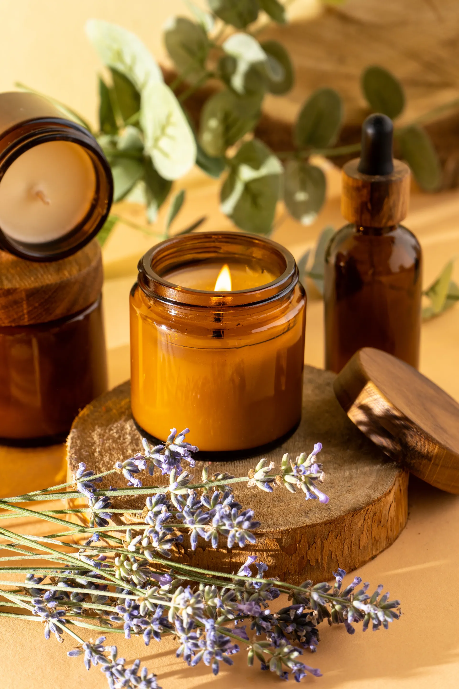 Lit candle in brown jar, lavender, essential oil bottle, on wooden coasters.
