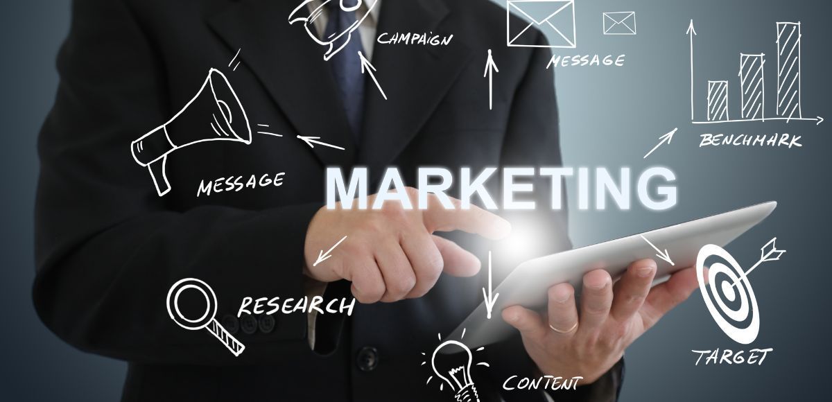 Hand tapping a tablet with “MARKETING” text and white icons on a dark background strategizing