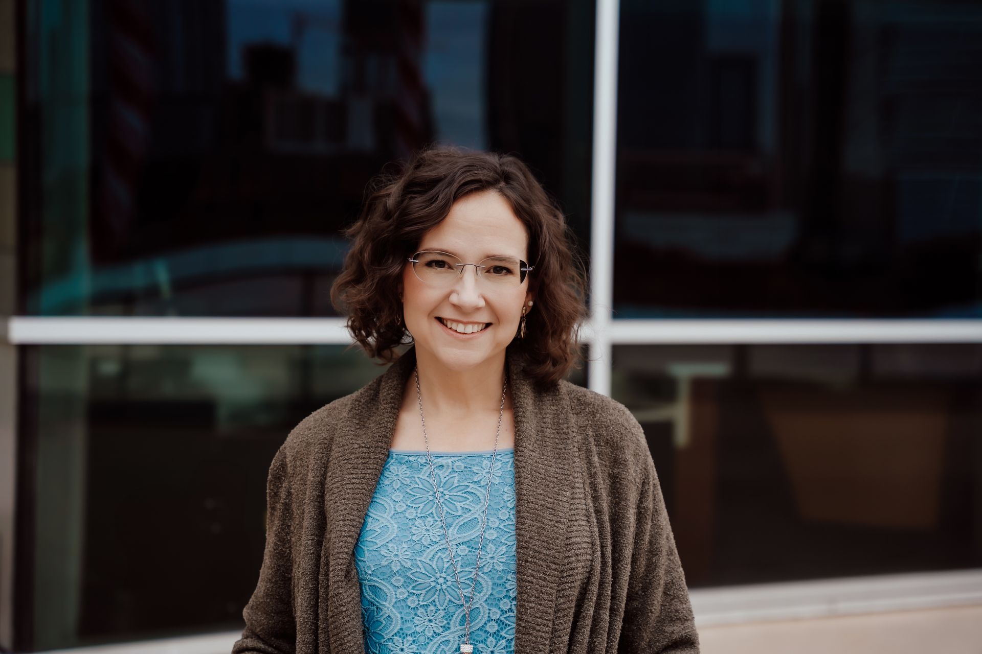 A woman wearing glasses and a brown sweater is standing in front of a building.