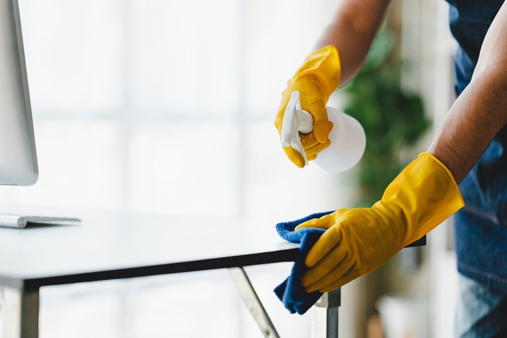 A person wearing yellow gloves is cleaning a desk with a spray bottle and a cloth.