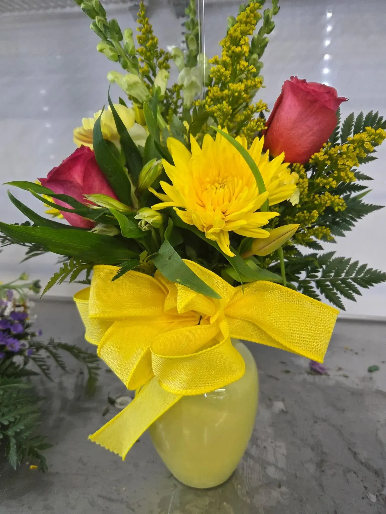 Yellow vase with yellow bow holding yellow daisy, two pink roses, and green foliage.