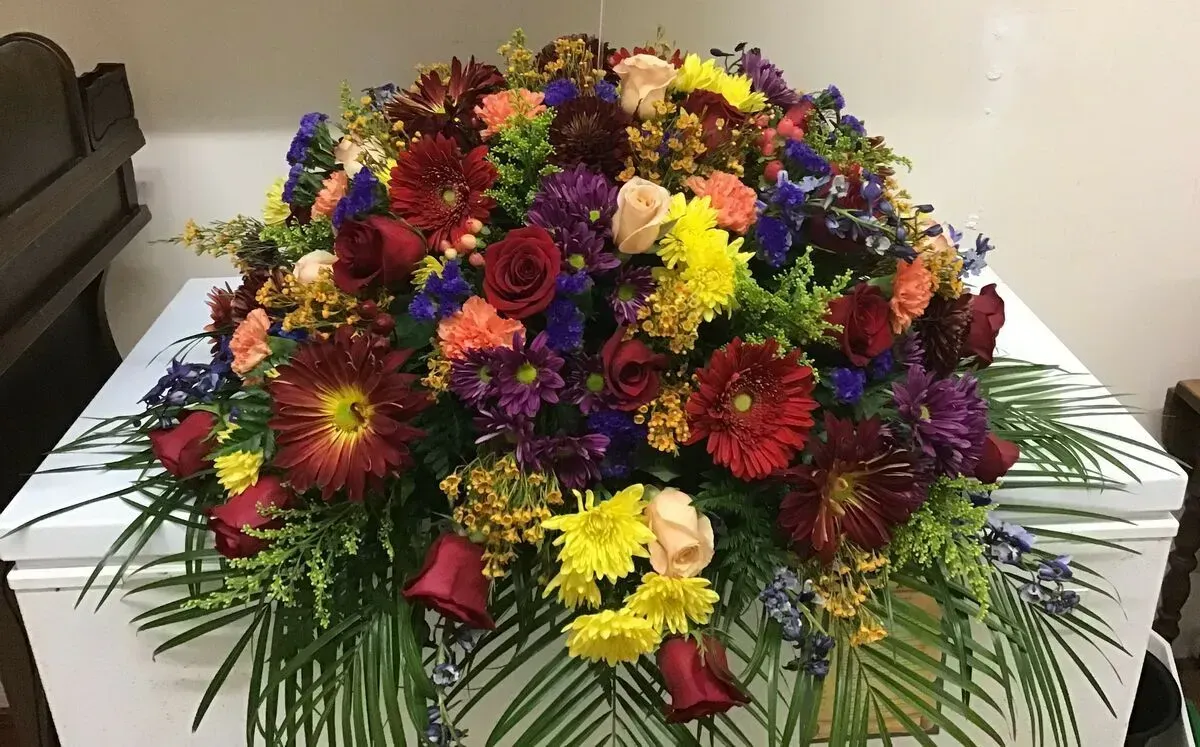 Casket covered in colorful floral arrangement: red, orange, yellow, and purple blooms with green foliage.
