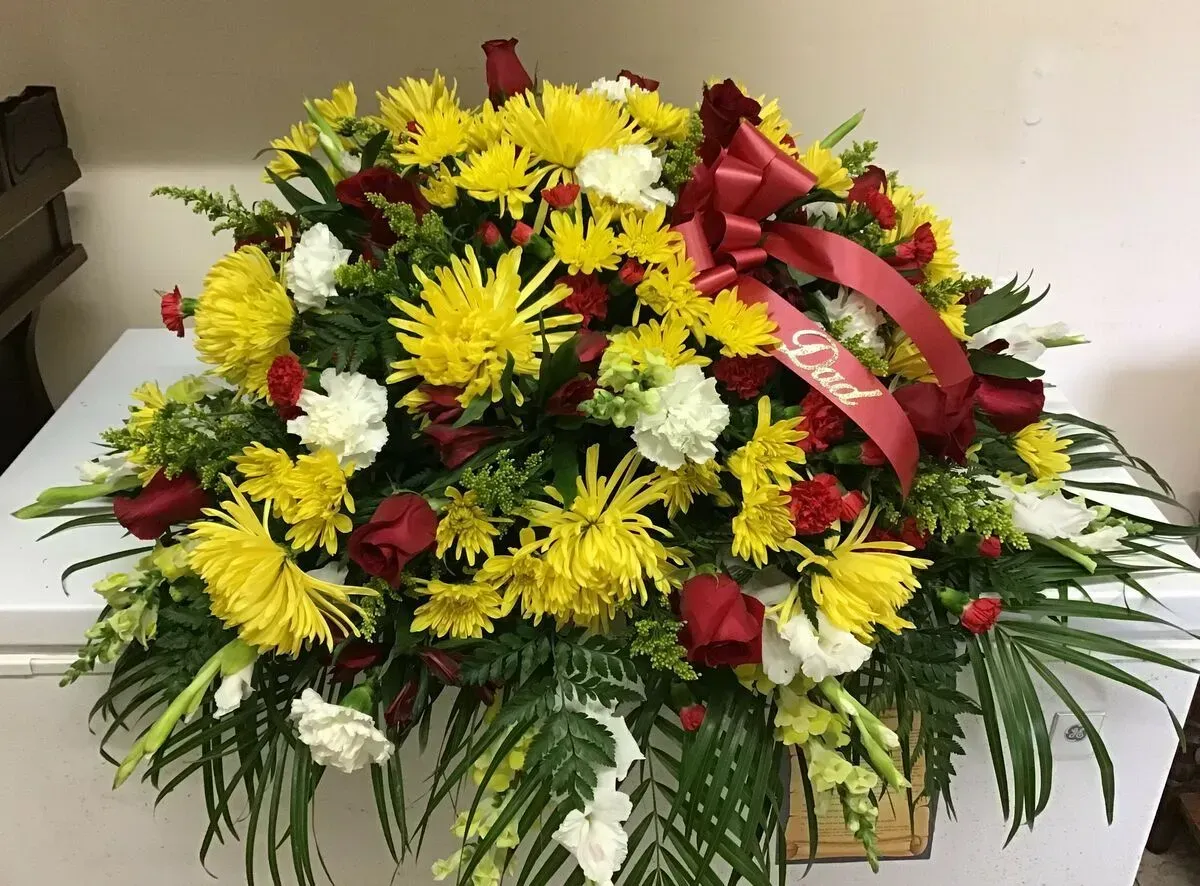 Funeral arrangement on a white surface, yellow and red flowers with a Dad ribbon.