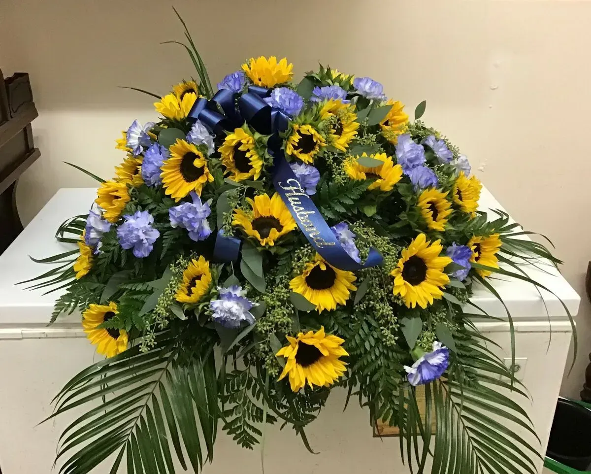 Funeral floral arrangement with sunflowers and blue flowers on a white surface.