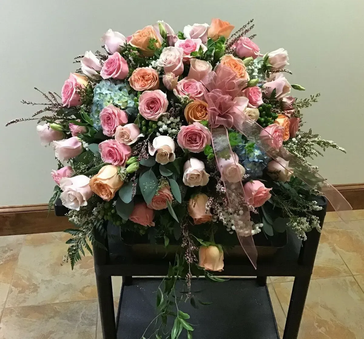 Floral arrangement with pink, peach, and white roses, greenery, and a ribbon, on a black stand.