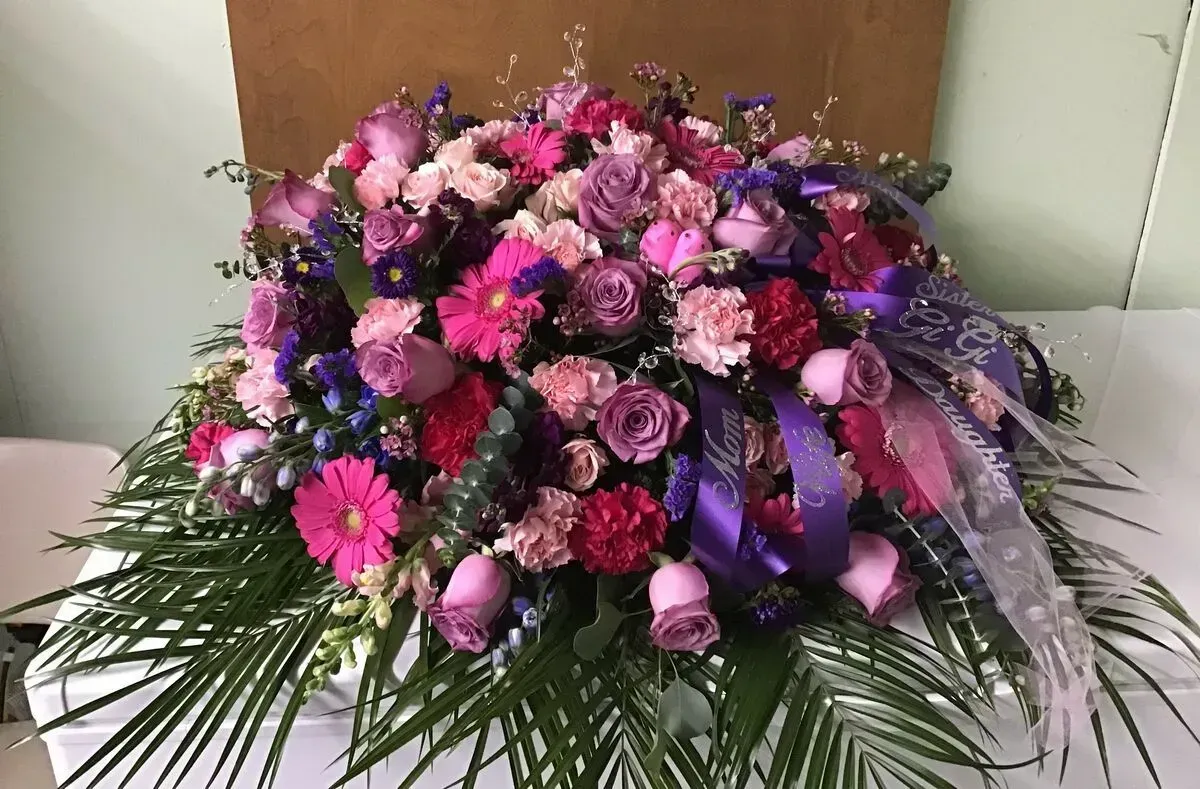 Floral casket spray with pink, purple, and red flowers, green foliage, and purple ribbon.