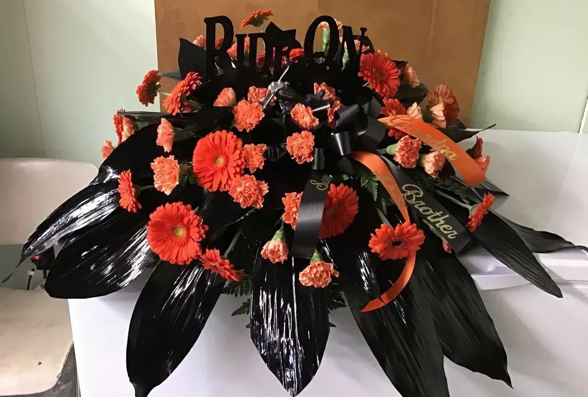 Floral arrangement with orange flowers, black leaves, and ribbon that says Reuben.