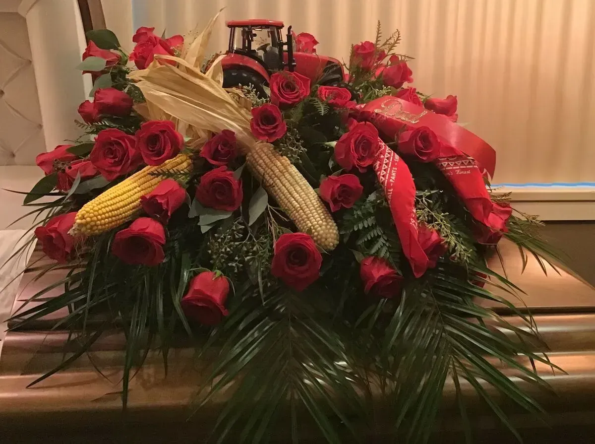 Floral arrangement with red roses, corn, and a toy tractor on a casket.