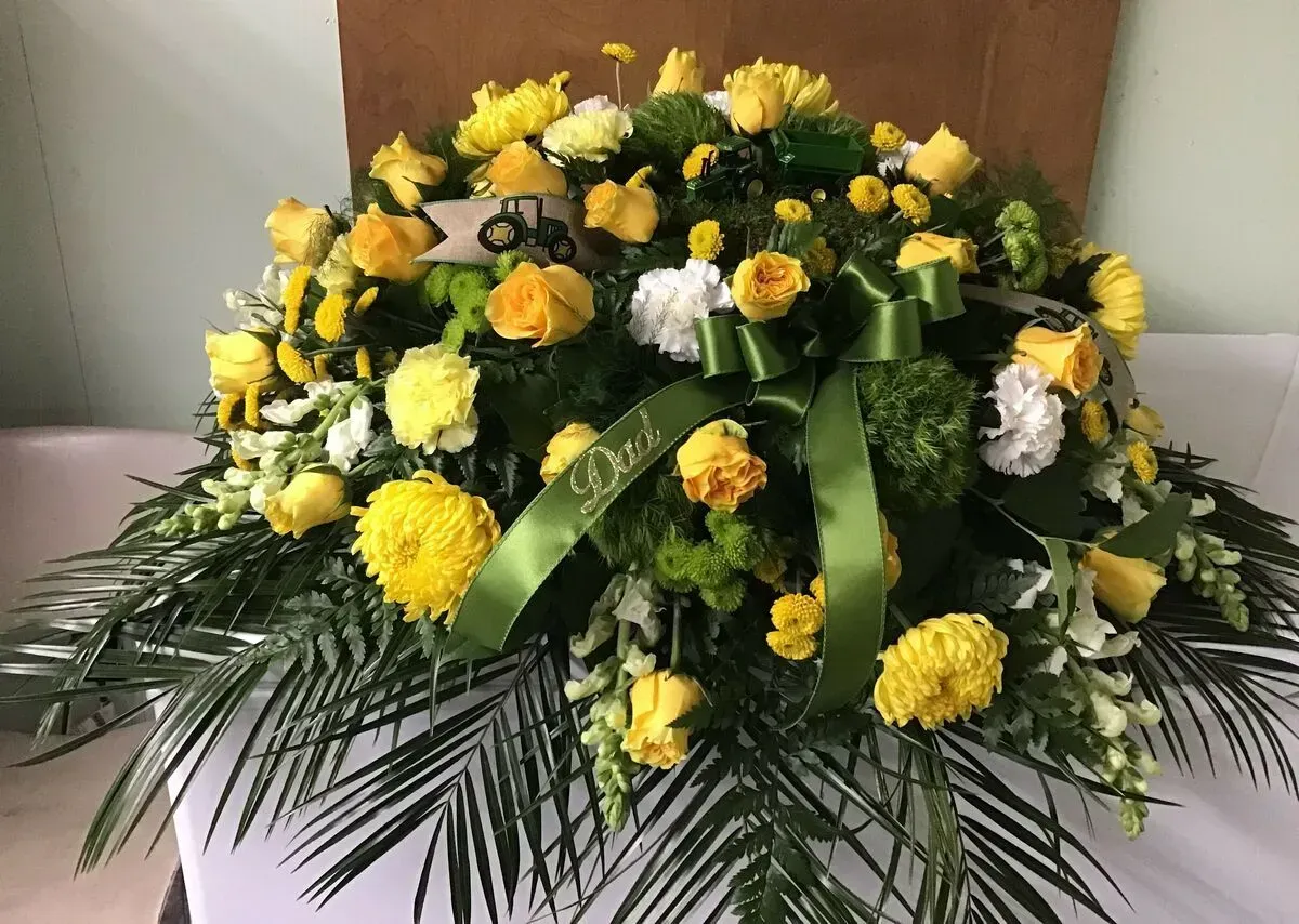 Floral arrangement, mostly yellow, green and white, on white surface. Green ribbon visible.