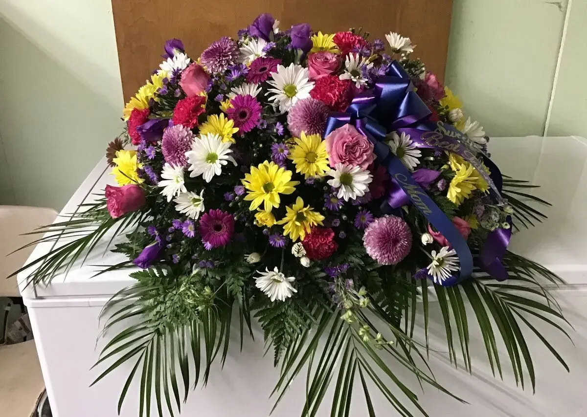 Floral casket spray with purple, yellow, and pink flowers, a blue ribbon, and green foliage.