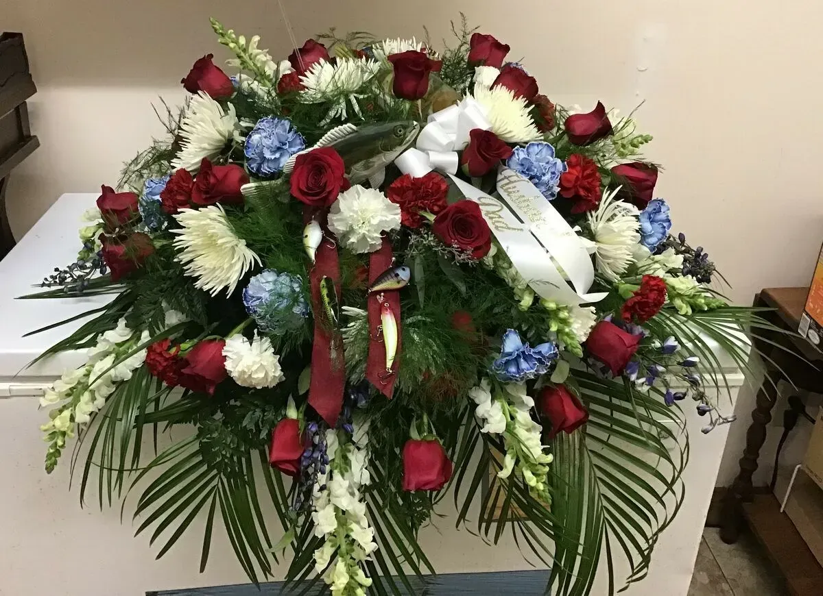 Casket spray of red roses, blue flowers, and white carnations with greenery and white ribbon.