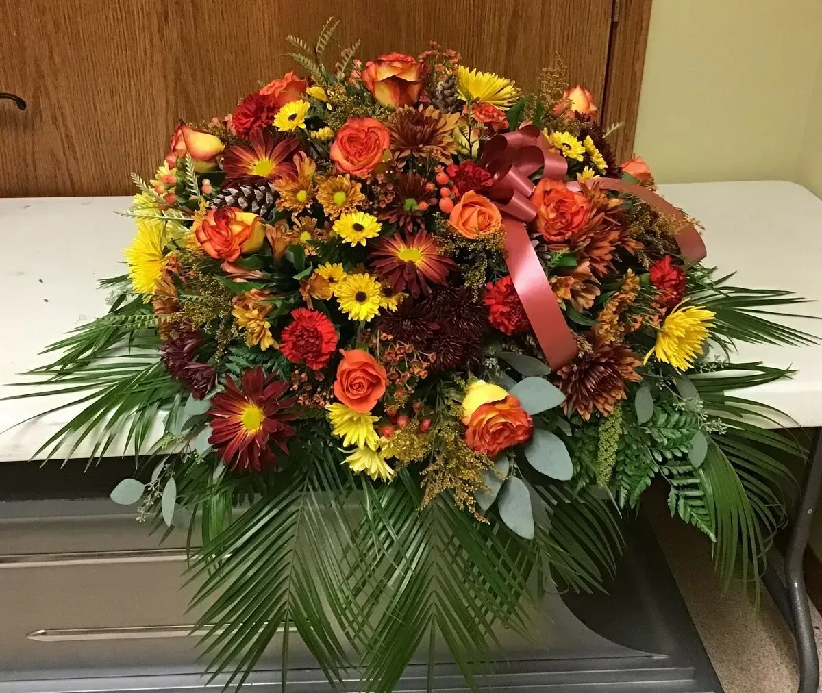 Floral casket spray with fall colors: orange, red, yellow blooms, greenery, and a maroon bow.