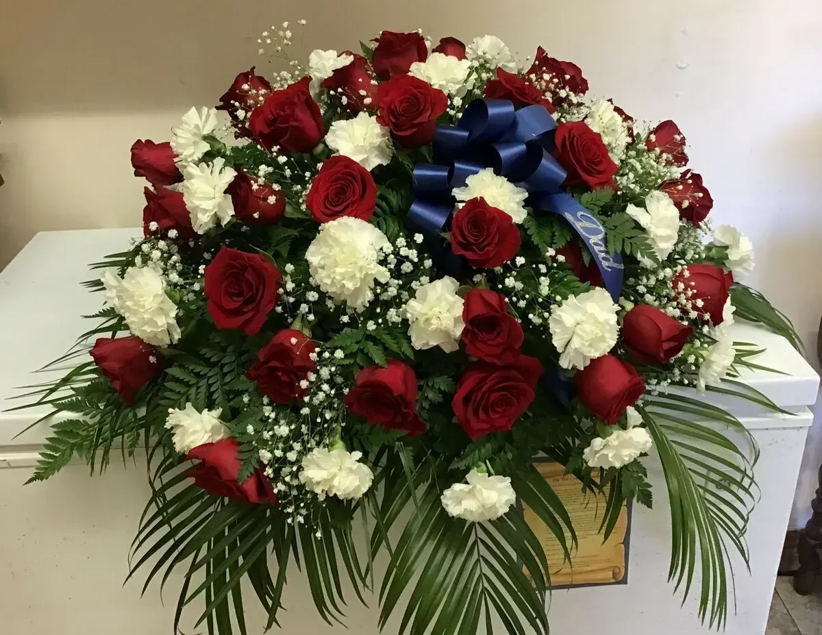 Funeral floral arrangement: red roses, white carnations, blue bow, greenery.