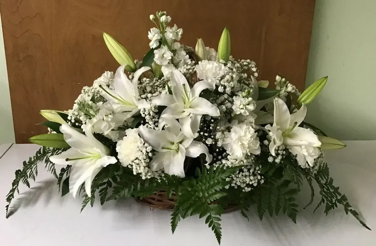 White floral arrangement in a woven basket, featuring lilies, carnations, and greenery.