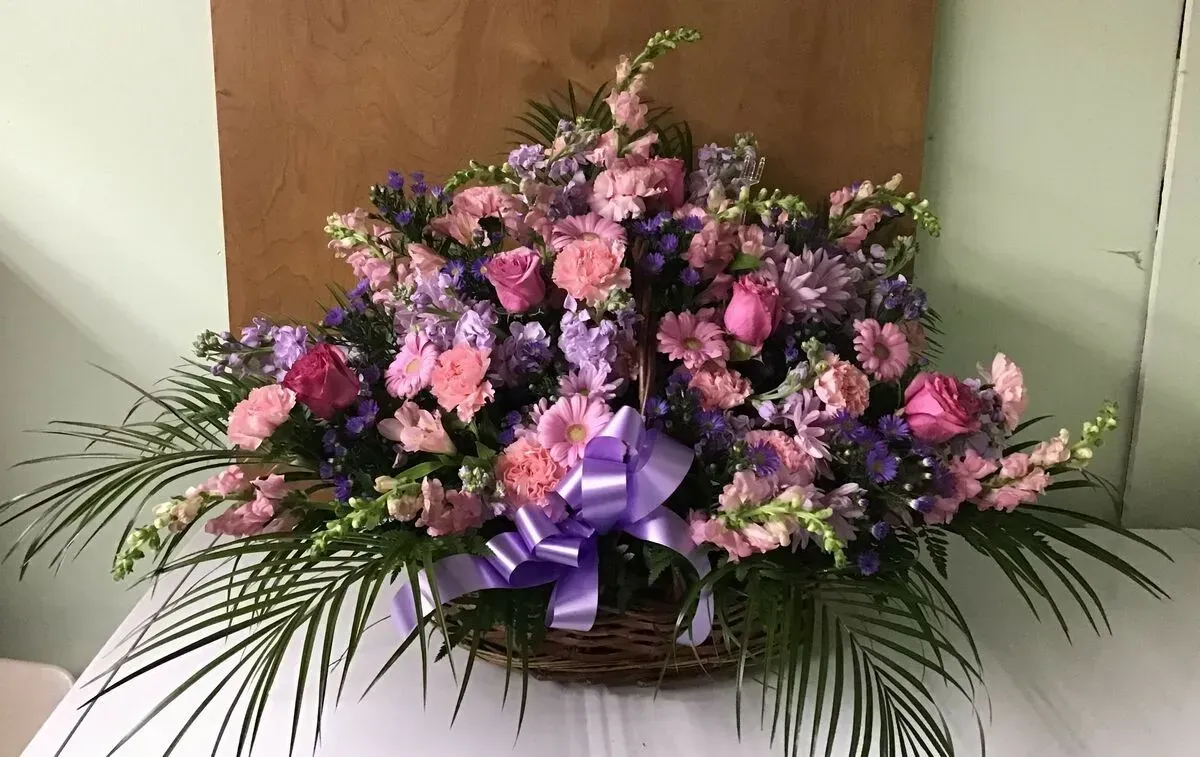 Floral arrangement in a woven basket, featuring pink, purple, and green flowers with a purple ribbon.