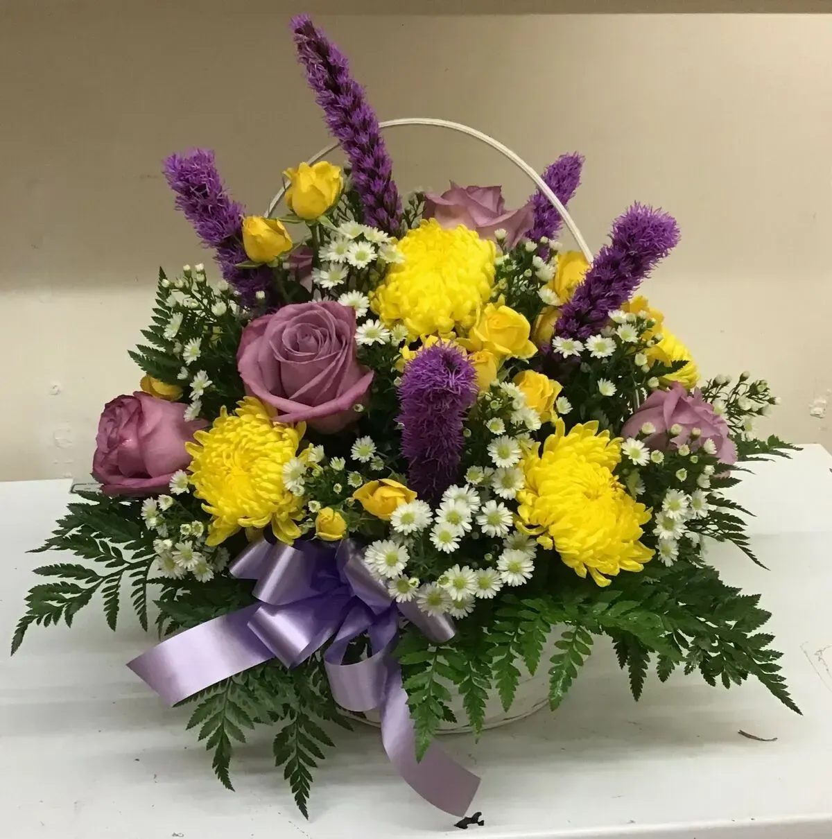 Floral arrangement in a white basket, with yellow and purple flowers, greenery, and a lavender bow.