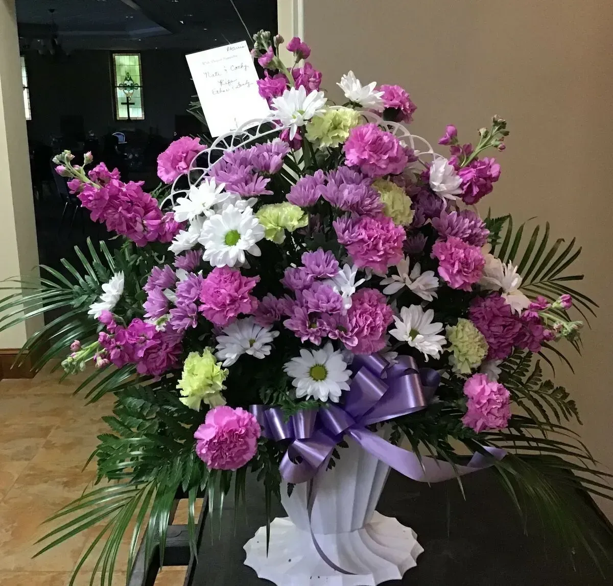 Floral arrangement in a white vase with purple carnations, daisies, and a lavender bow.