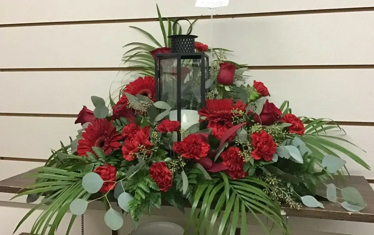 Floral arrangement with red flowers, green foliage, and a black lantern on a wooden shelf.