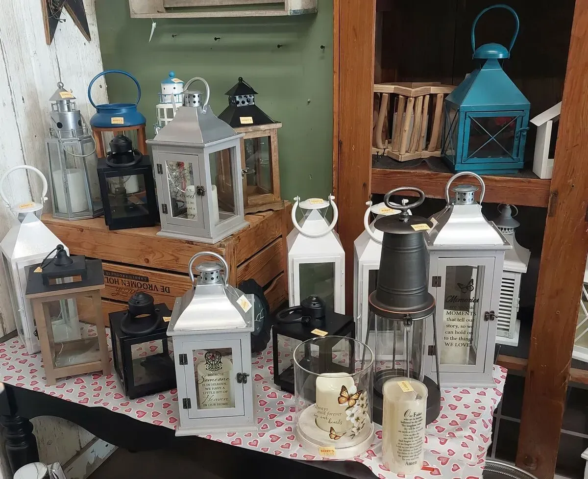 Display of various lanterns on a table, including white, black, and teal, with wooden shelving in the background.