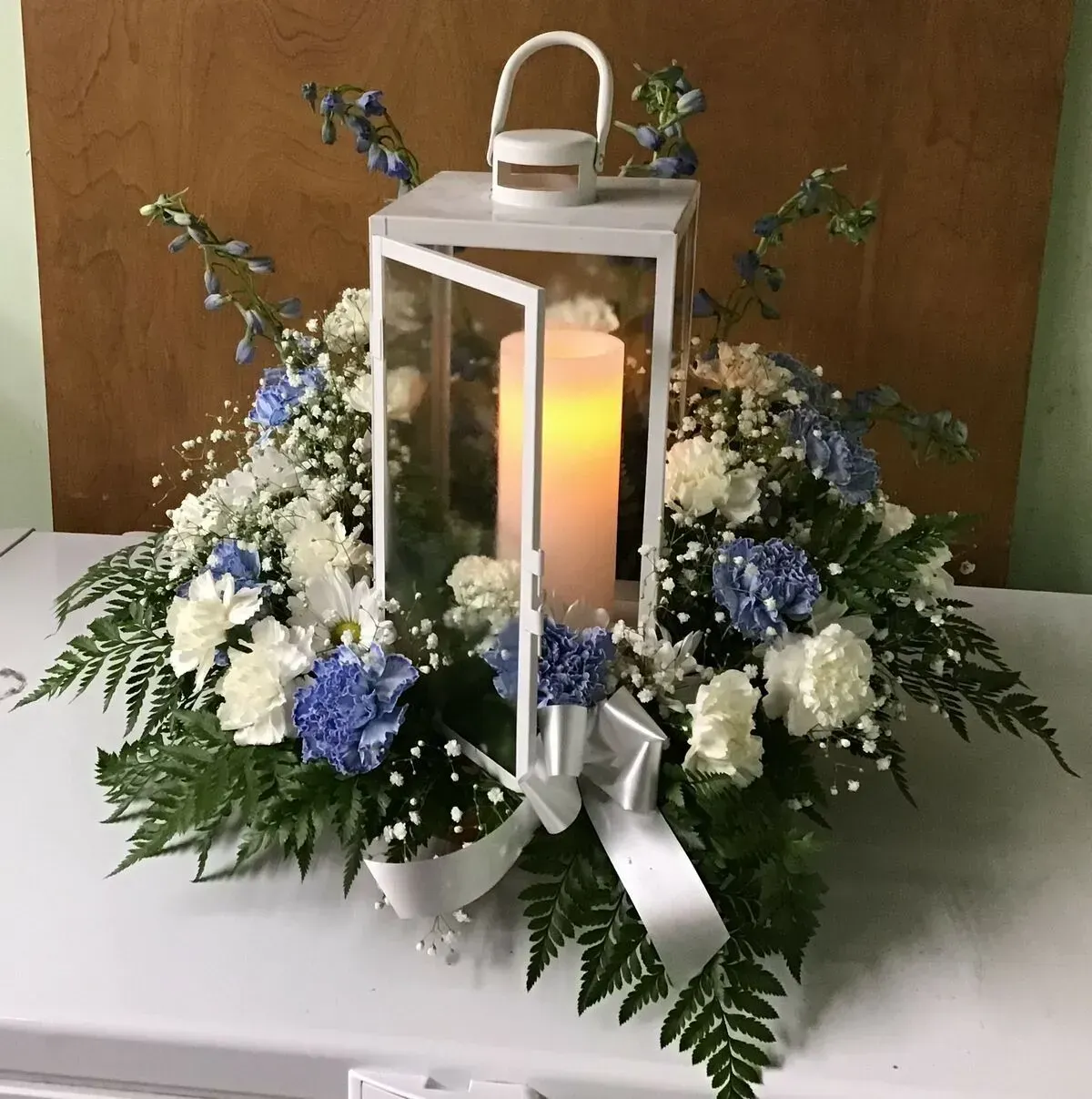 White lantern with lit candle, surrounded by blue and white flowers and greenery.