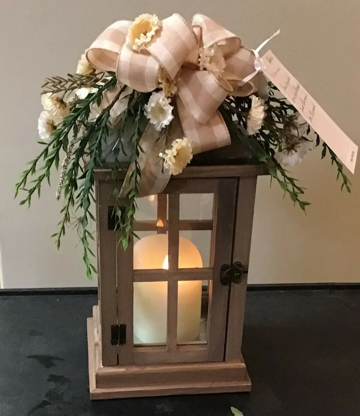 Wooden lantern with lit candle, decorated with flowers and a plaid bow.