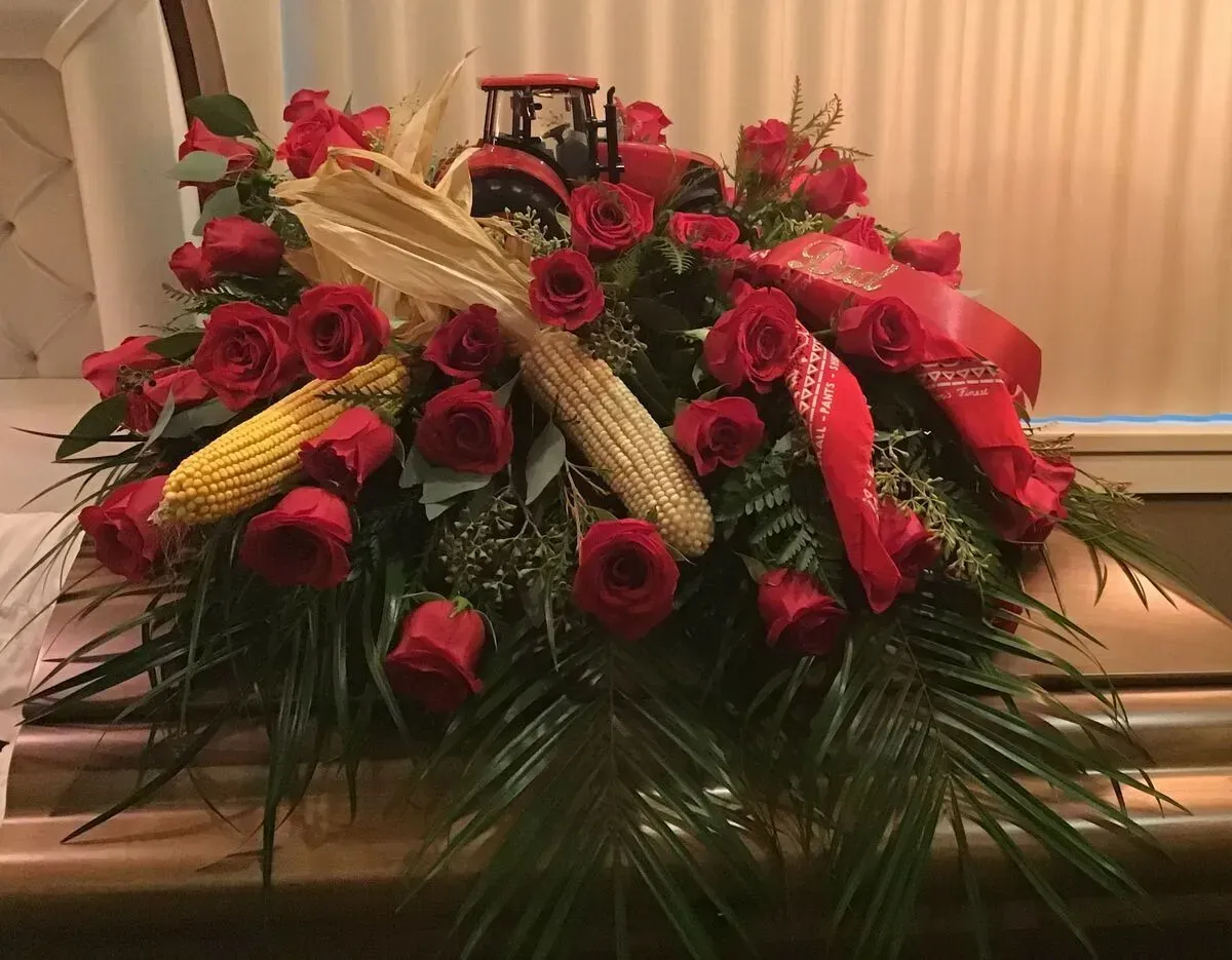 Red roses, corn, and a toy tractor adorn a casket, with greenery and red ribbon.