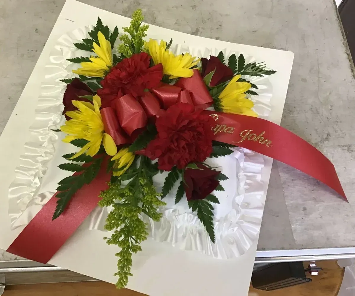 Floral arrangement with red, yellow, and green flowers on a white square base with red ribbon.