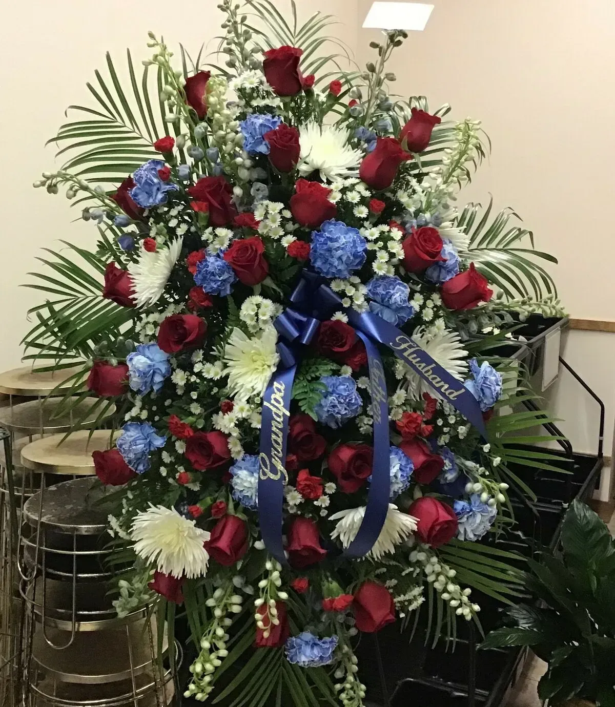 Floral standing spray with red roses, blue flowers, white chrysanthemums, and a blue ribbon.