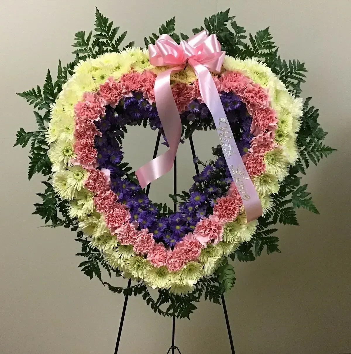 Heart-shaped floral arrangement on a stand with pink ribbon, cream and pink flowers, and ferns.