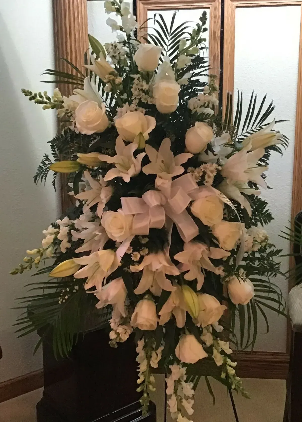 White floral arrangement on a stand, possibly for a memorial; features roses, lilies, and a bow.