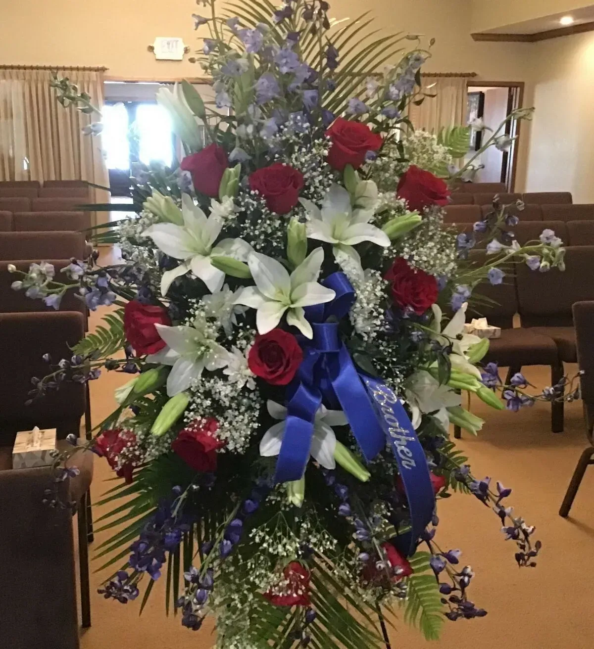 Floral arrangement with red roses, white lilies, and blue accents, ribbon reads Brother, inside a chapel.