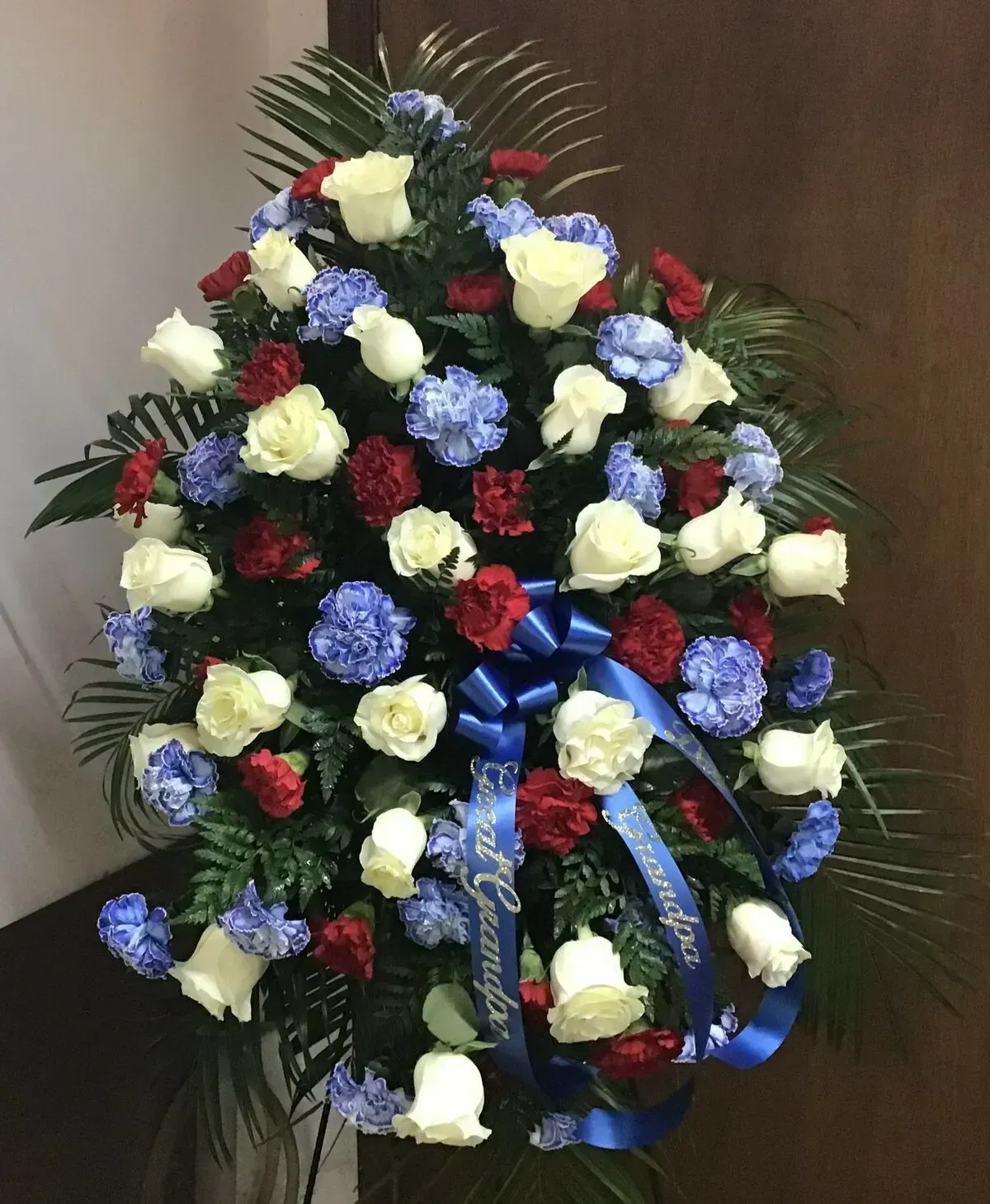 Standing floral arrangement with white roses, blue carnations, and red carnations, with a blue ribbon.