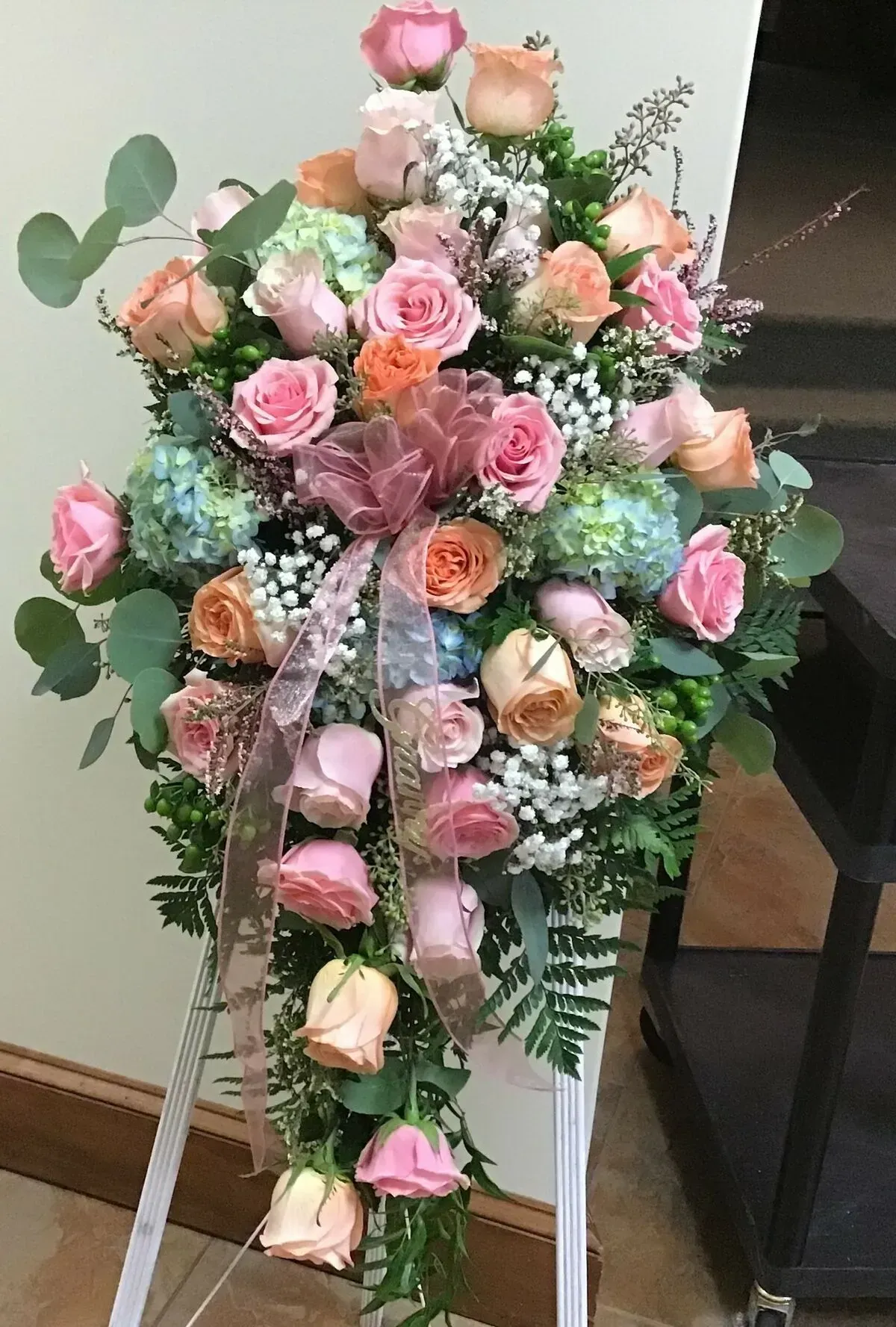A floral spray of peach and pink roses, hydrangeas, and greenery on a white easel.