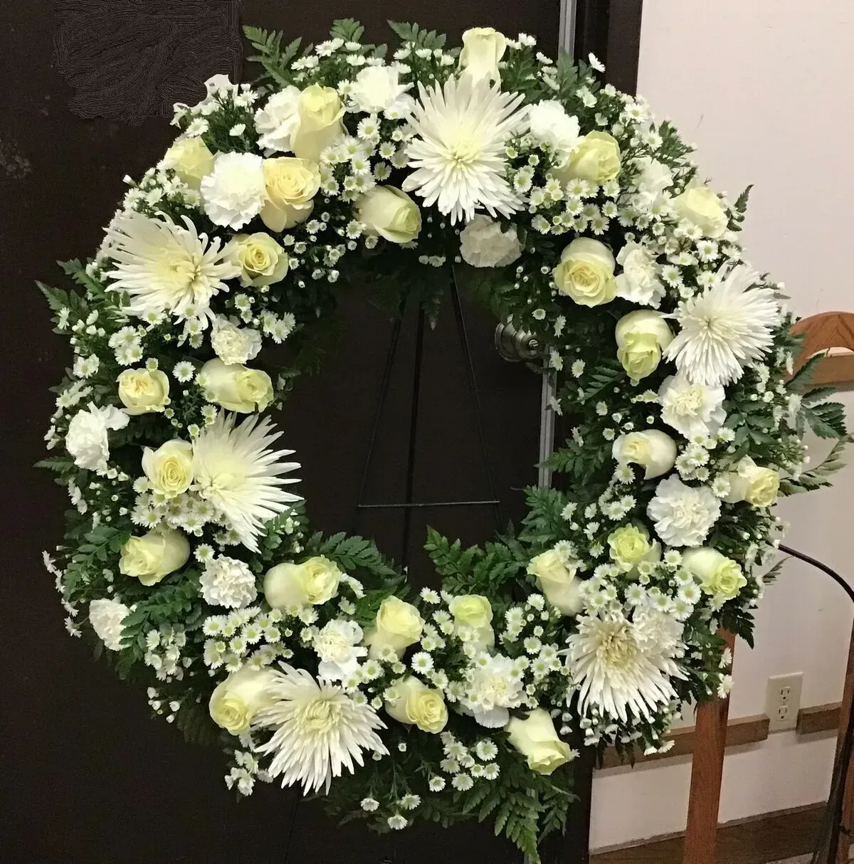White floral wreath with roses and mums on a stand, likely a memorial arrangement.