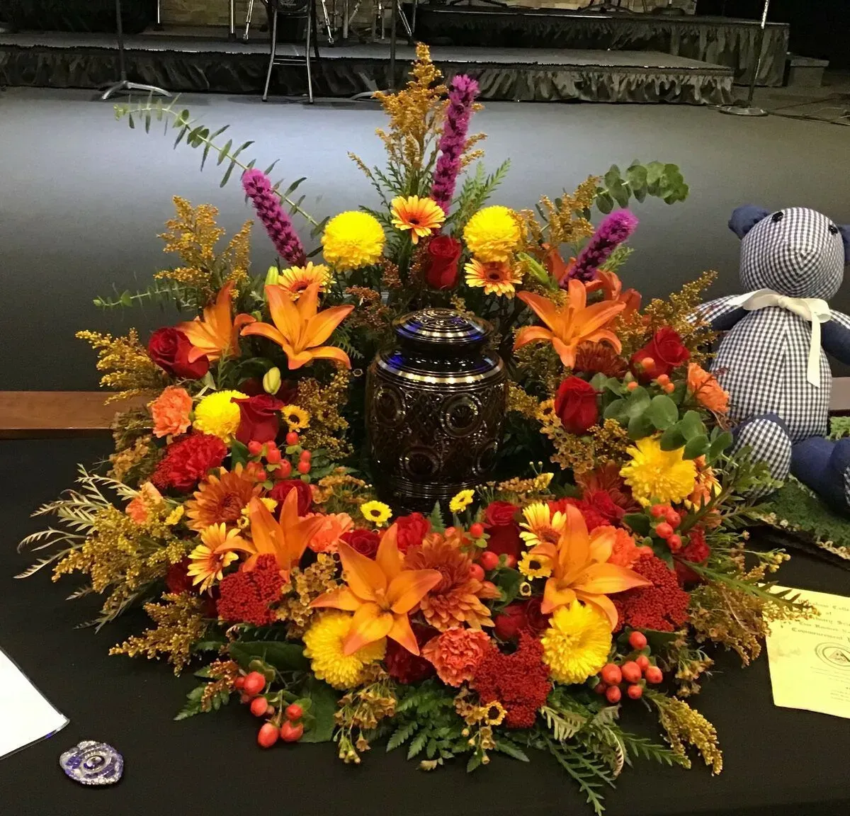 Floral arrangement surrounding a cremation urn; orange lilies, yellow daisies, red carnations, and purple accents.