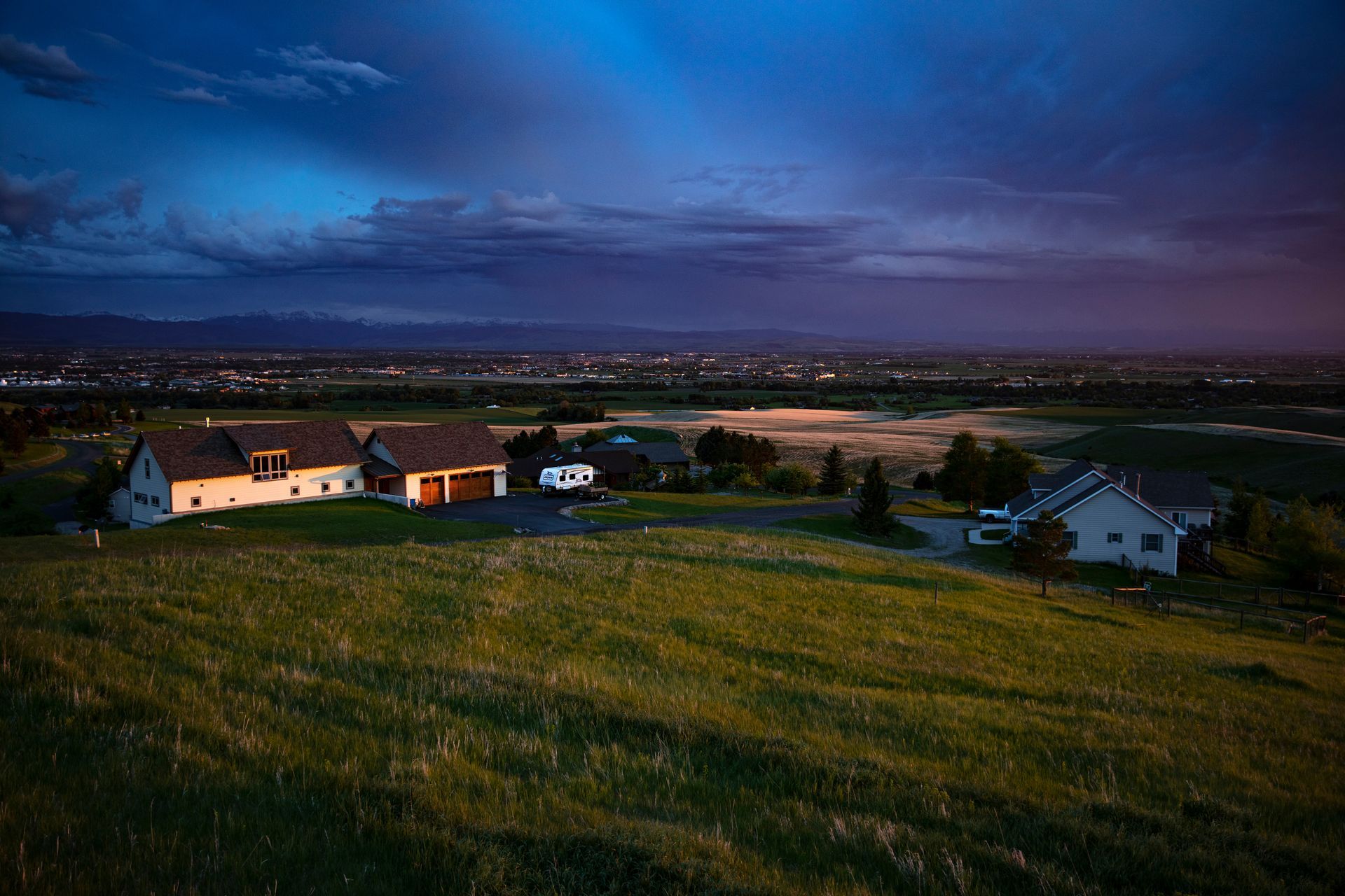 Houses on a grassy hill at dusk, city lights in the distance under a blue and purple sky. Houses on a grassy hill at dusk, city lights in the distance under a blue and purple sky.