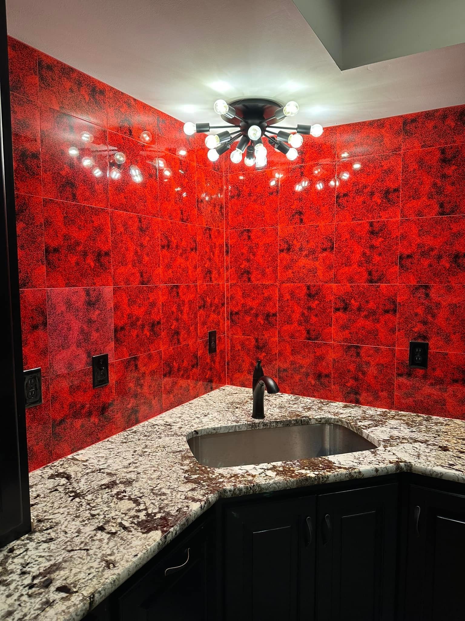 A modern corner wet bar featuring high-contrast red and black patterned wall tiles, granite countertops, and black cabinets.