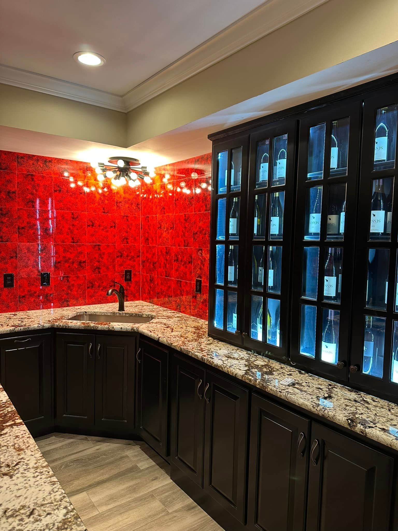 A bar with dark wooden cabinets, granite countertops, a corner sink, and a vibrant, textured red tile backsplash.