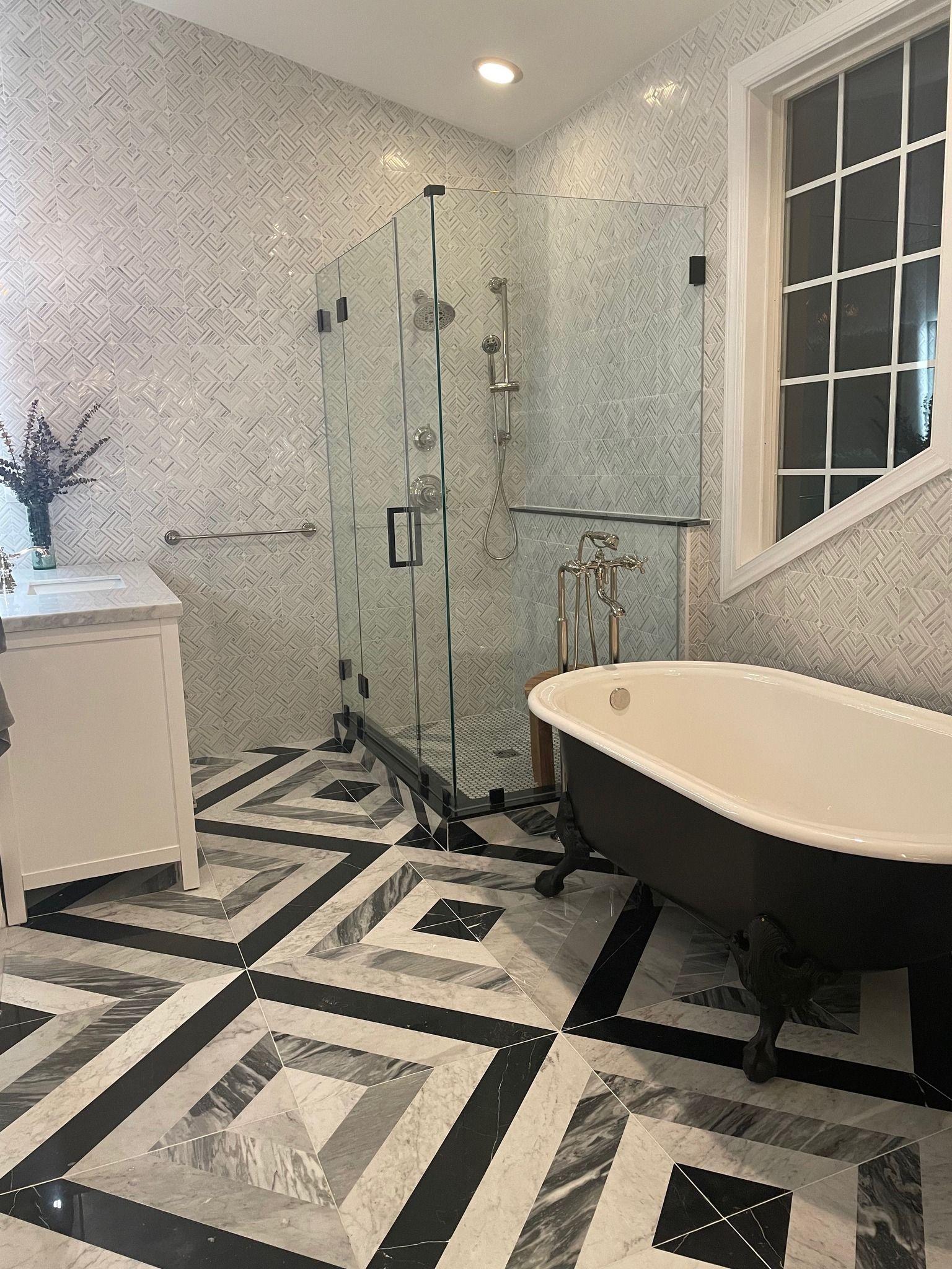 A bright bathroom with a black clawfoot tub, glass walk-in shower, marble walls, and a geometric black and white tile floor.