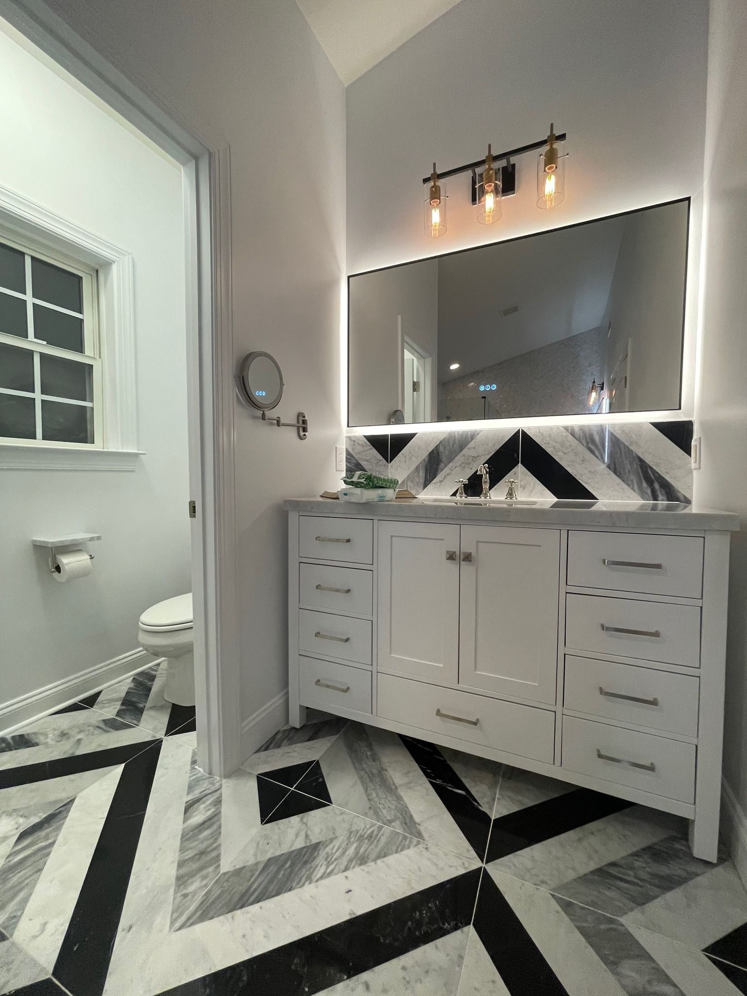 A white vanity with a large backlit mirror and chevron-patterned marble flooring in a bathroom with a separate toilet.
