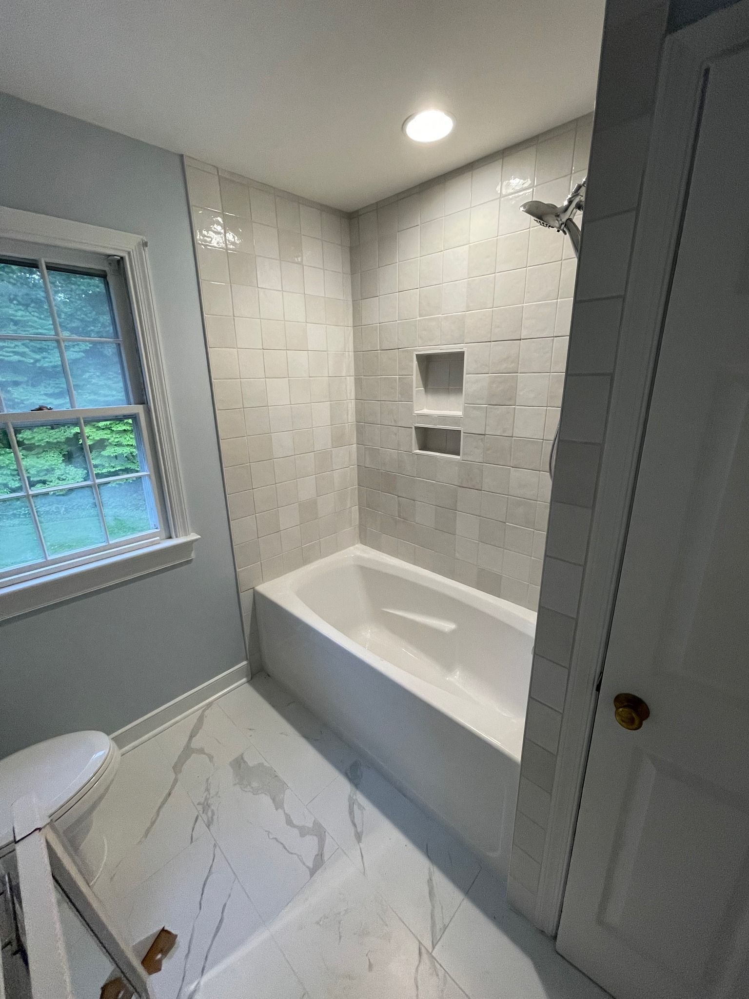 A bathroom featuring a white bathtub with a tile surround, built-in shelving, white marble flooring, and a window.