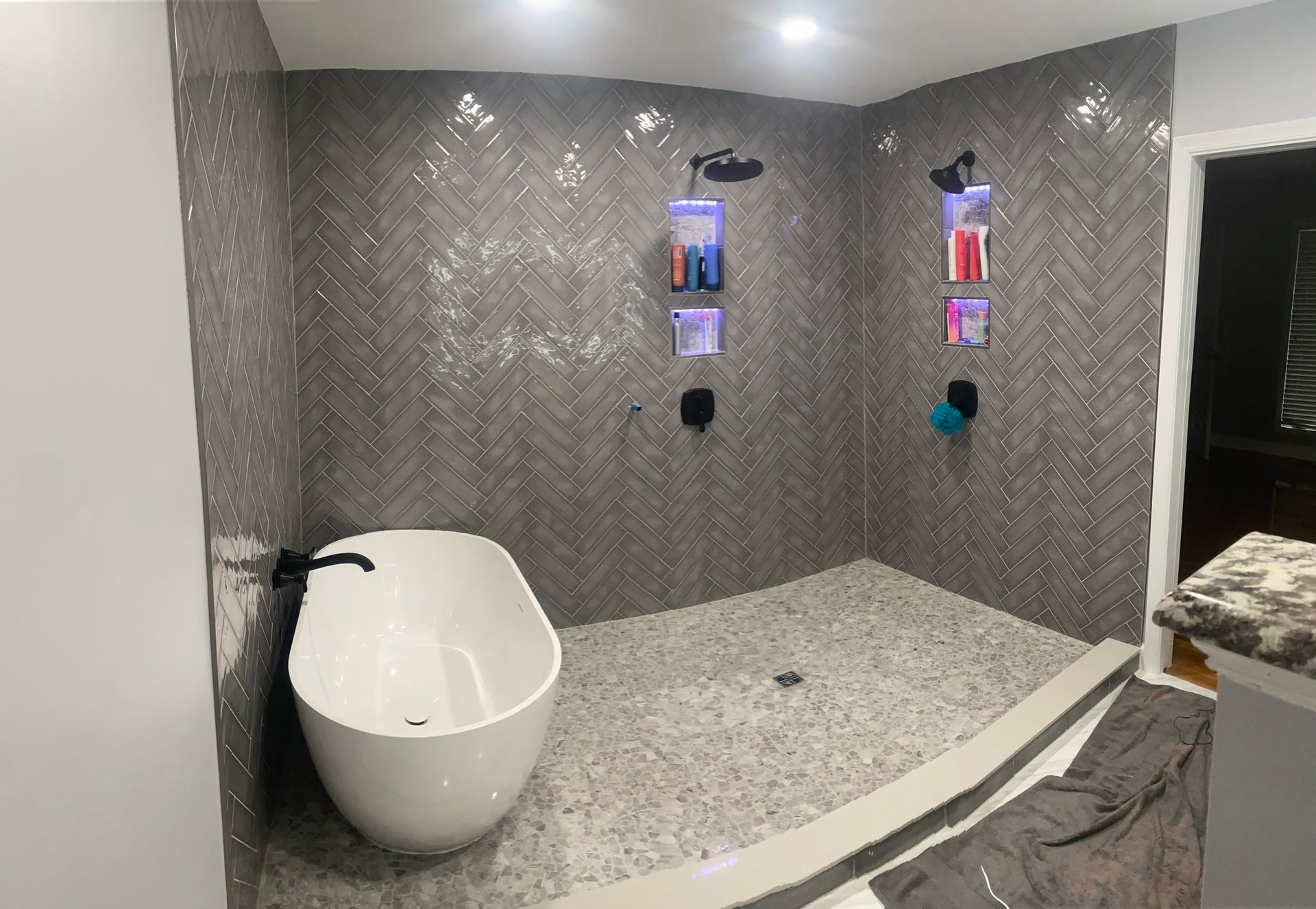 Modern bathroom with a freestanding white tub next to a walk-in shower featuring grey herringbone tiles and pebble floors.